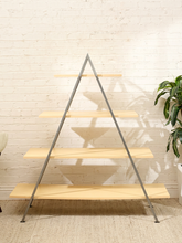 Load image into Gallery viewer, A modern A-frame shelving unit with five light natural wood shelves and a dark gray metal frame, set against a white brick wall with a plant on the right.