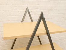 Load image into Gallery viewer, A close-up of a modern shelving unit or table with light natural wood panels and dark gray metal A-frame legs, set against a white painted brick wall.