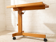 Load image into Gallery viewer, A light natural wood rolling table with a folding top, adjustable central support, and black caster wheels, set against a white brick wall and light carpet.
