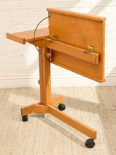 Load image into Gallery viewer, A light natural wood adjustable lecture stand with a writing surface, side table, and caster wheels, shown against a white brick wall and beige rug.