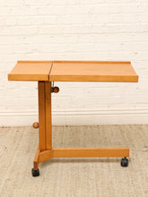 Load image into Gallery viewer, A mid-century modern wooden rolling table with a light natural wood finish, featuring a split tabletop, adjustable height knobs, and black caster wheels, set against a white brick wall and beige rug.