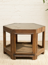 Load image into Gallery viewer, A hexagonal wooden side table with a dark brown finish, featuring a top surface with a light stone inlay and an open lower shelf, set against a white brick wall and beige rug.