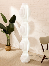 Load image into Gallery viewer, A tall, modern white floor lamp with a sculptural, wavy ribbed design stands in a room corner against a white brick wall. A potted green plant is on the left, and a light-colored chair is on the right.
