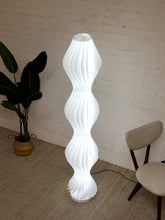 Load image into Gallery viewer, A modern white spiral floor lamp with a ribbed, wave-like design stands in a room corner against a white brick wall, next to a potted plant and a light-colored chair.