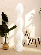 Load image into Gallery viewer, A tall, sculptural white floor lamp with a ribbed, wavy design stands in a bright room next to a potted plant and a wooden chair.