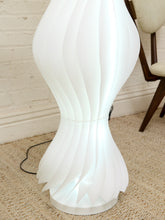 Load image into Gallery viewer, A modern white floor lamp with a sculptural, wavy ribbed design, illuminated from within to cast a soft glow. It is shown standing on a beige textured carpet against a white brick wall, with a wooden chair leg visible in the background.