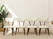 Load image into Gallery viewer, Four mid-century modern dining chairs with cream upholstered seats and curved wooden backs, arranged in a row against a white brick wall with a potted plant on the left.