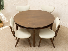 Load image into Gallery viewer, A round mid-century modern dining table with a dark brown wood finish, surrounded by four matching chairs with cream upholstery and angled dark wood legs, set on a beige carpet against a white brick wall.
