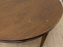 Load image into Gallery viewer, A close-up of a round wooden table top with visible wear, scratches, and chipped paint on the brown wood grain, resting on a beige woven rug.
