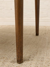 Load image into Gallery viewer, Close-up of a dark brown wooden table leg standing on a textured beige woven rug in front of a white brick wall.