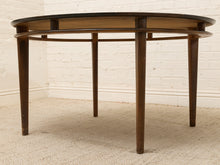 Load image into Gallery viewer, A round mid-century modern wooden coffee table with a dark top, warm brown frame, and tapered legs, set against a white brick wall and beige carpet.