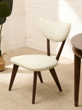 Load image into Gallery viewer, A mid-century modern dining chair with a cream upholstered seat and curved backrest, supported by dark brown wooden legs, shown in a bright room next to a table.