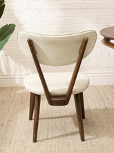 Load image into Gallery viewer, A mid-century modern dining chair with a cream-colored upholstered seat and curved backrest, supported by a dark brown wooden frame with tapered legs, set against a white brick wall and light beige rug.