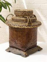 Load image into Gallery viewer, A traditional hand-woven bamboo storage jar with a matching lid, resting on a dark wooden base. The jar features a dark brown woven body, a lighter tan woven neck and handles, and a wooden stand with small brass corner accents.