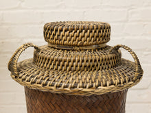 Load image into Gallery viewer, A woven natural fiber basket with a lid, featuring two side handles and a smaller round lid stacked on top, set against a white painted brick wall background.
