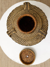 Load image into Gallery viewer, A top-down view of a round woven basket with a matching lid, displayed on a white circular surface against a beige textured background.