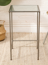 Load image into Gallery viewer, A square bar-height side table with a clear glass top and a weathered gray metal frame, set on a woven jute rug against a white brick wall.
