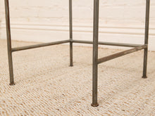 Load image into Gallery viewer, Close-up of a rustic metal side table leg frame with weathered dark gray iron tubes, standing on a beige woven jute rug against a white brick wall.