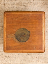 Load image into Gallery viewer, A square wooden board or tray with rounded corners and a circular dark metal emblem in the center, photographed from above on a woven beige fabric background.