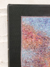 Load image into Gallery viewer, Close-up of a framed textile art piece showing a textured black frame and a colorful woven pattern in shades of blue, pink, and orange against a white brick wall background.