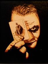 Load image into Gallery viewer, A close-up artwork of a Joker face made from wood burning or engraving, holding a real Joker playing card in front of one eye against a black background.
