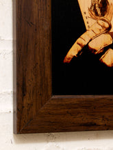 Load image into Gallery viewer, Close-up of a framed artwork showing a dark wood frame corner against a white brick wall, with a black background and part of a warm-toned painted figure visible.