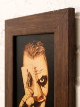 Load image into Gallery viewer, A close-up of a framed artwork showing a stylized portrait of a face with dramatic eye makeup and a playing card covering one eye, mounted against a light textured wall.