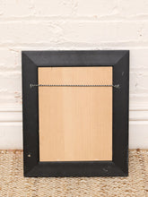 Load image into Gallery viewer, The back of a black picture frame with a light wood backing board and a hanging wire, resting on a woven rug against a white brick wall.