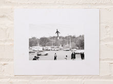 Load image into Gallery viewer, Vintage black-and-white photograph of a public park or plaza with people, period vehicles, trees, and a tall central structure on a white mount