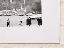 Load image into Gallery viewer, A black and white photographic print mounted on a white board, depicting a vintage street scene with buses, cars, and several people standing outdoors.