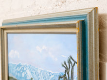 Load image into Gallery viewer, A close-up of a framed landscape painting showing blue mountains and a Joshua tree under a pale sky, set in a distressed teal and beige frame against a white brick wall.