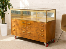 Load image into Gallery viewer, A vintage-style wooden display case with six drawers, brass handles, a glass top, and red caster wheels, photographed in a bright room with a white brick wall, potted plant, and chair.