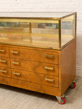 Load image into Gallery viewer, A vintage mid-century modern rolling display cabinet with a burl wood finish, brass hardware, and a glass display case on top, set against a white brick wall.