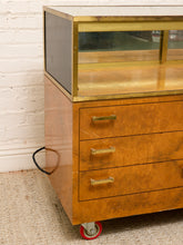 Load image into Gallery viewer, A vintage mid-century wooden display cabinet with three drawers, brass handles, a glass top display case, and red caster wheels, photographed in an indoor setting against a white brick wall.