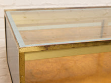 Load image into Gallery viewer, Close-up of a vintage display case or cabinet with a clear glass top, gold-toned metal frame, and warm amber-brown interior panels, set against a white brick wall.