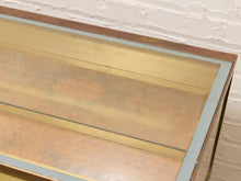 Load image into Gallery viewer, Close-up of a glass-top cabinet or display case with a warm wood frame and pale blue-gray trim, photographed against a white painted brick wall.