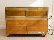 Load image into Gallery viewer, A vintage rolling storage cabinet with a warm orange-brown burl wood front, a brass top frame, and red caster wheels, set against a white brick wall.