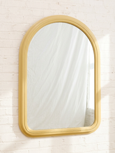 Load image into Gallery viewer, An arched wall mirror with a thick, rounded gold frame, mounted on a white painted brick wall.