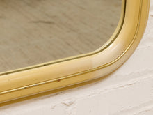Load image into Gallery viewer, Close-up of a vintage-style wall mirror with a curved, glossy cream and gold frame mounted on a white painted brick wall.