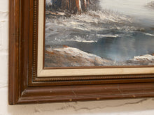 Load image into Gallery viewer, A close-up of a framed landscape painting showing a snowy shoreline, dark trees, and blue-gray water, with a dark brown wooden frame and cream mat against a white brick wall.
