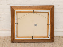 Load image into Gallery viewer, The back of a framed artwork, showing a medium brown wooden frame, cream canvas backing, white paper margins, and metal hanging hardware, set against a white brick wall and woven rug.