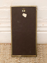 Load image into Gallery viewer, The back of a rectangular framed picture with a dark brown backing board and a brass frame, standing on a woven rug against a white brick wall.