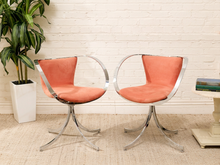 Load image into Gallery viewer, Two mid-century modern swivel chairs with curved chrome metal frames and coral orange upholstered seats, shown in a styled room setting with a potted plant and a side table.