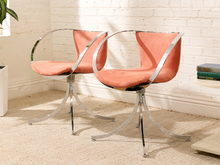 Load image into Gallery viewer, Two modern swivel dining chairs with curved coral pink fabric seats and backs, supported by chrome-finished tubular steel frames and curved splayed legs, set in a room with a white brick wall and beige carpet.