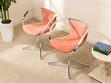 Load image into Gallery viewer, Two modern swivel accent chairs with peach-pink velvet upholstery and curved chrome metal frames, shown in a styled room setting with a white brick wall, beige carpet, and a side table.