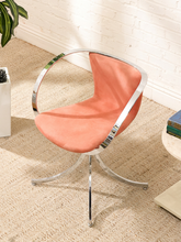 Load image into Gallery viewer, A modern accent chair with a curved, upholstered peach-orange seat and backrest, supported by a chrome-finished swivel base and curved armrests, set on a woven rug against a white brick wall.