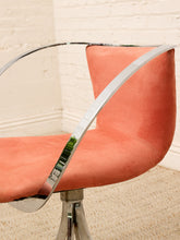 Load image into Gallery viewer, A close-up of a modern swivel chair with a curved, upholstered seat in a warm coral or salmon pink fabric, supported by a polished chrome metal frame and base, set against a white brick wall and beige carpet.