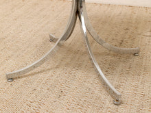 Load image into Gallery viewer, Close-up of a polished chrome table base with three curved legs, shown on a beige woven jute rug.