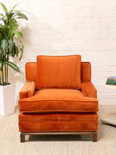 Load image into Gallery viewer, An orange velvet armchair with a matching back cushion, set against a white brick wall background with a potted plant on the left and a small side table on the right.