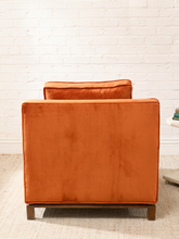 Load image into Gallery viewer, An orange velvet armchair with a wooden base, photographed from the back against a white brick wall and light beige rug.